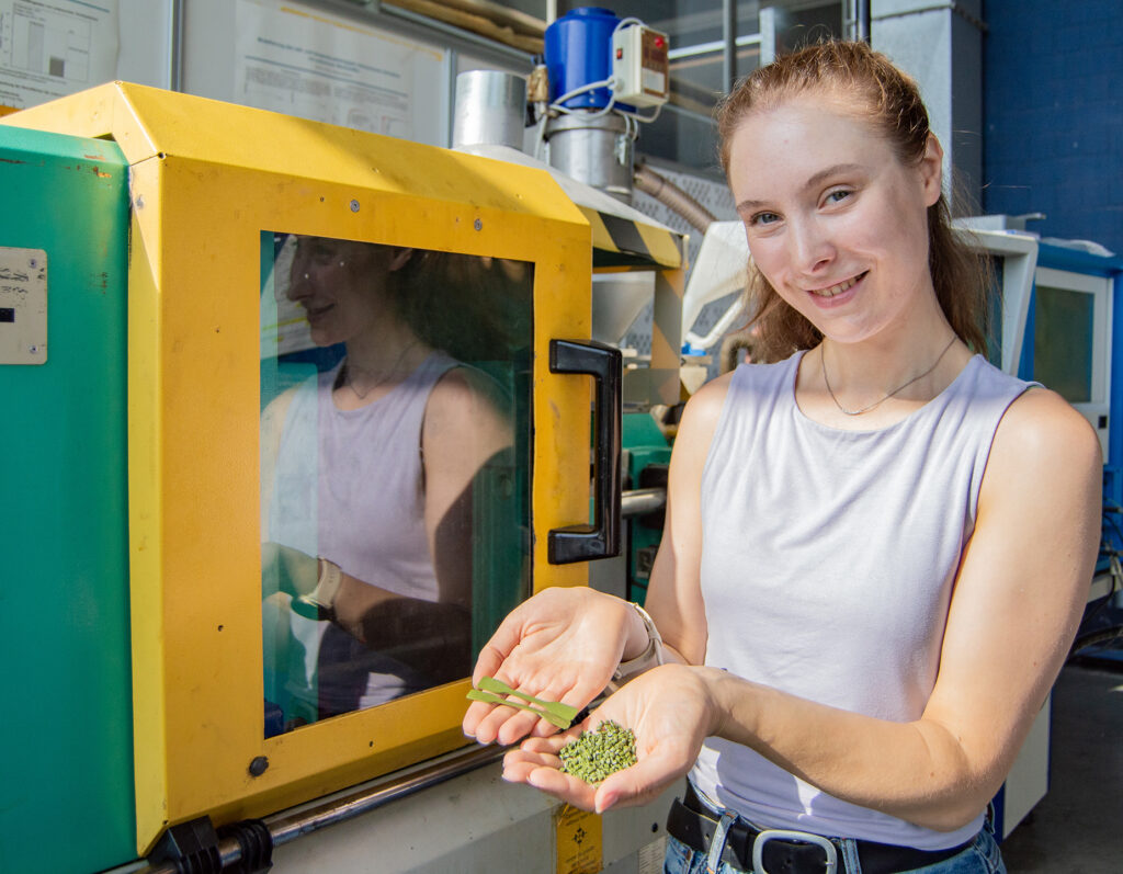 Doktorandin Anna lena Seibel mit ersten Ergebnissen aus dem Forschungsprojekt RewitAl der Arbeitsgruppe von Professorin Taha an der Hochschule Aalen. Die 27-Jährige präsentiert grünes Granulat, das aus altem Kunststoffrasen gewonnen wurde, vor einer Kunststoffspritzgussmaschine.